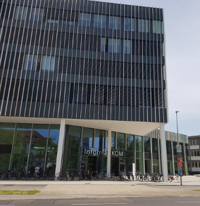 Das InformatiKOM des Karlsruher Instituts für Technologie ist zu sehen. Man sieht das Bild eines Gebäudes. Das Gebäude ist im unteren Bereich von einer gläsernen Front und im oberen Bereich von einer grauen Fassade durchzogen. Vor dem Gebäude stehen mehrere Fahrräder und eine Straße ist zu sehen.