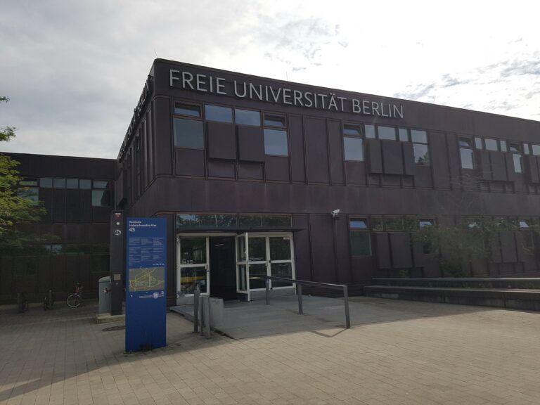 Das Bild zeigt die Frontseite eines Gebäudes mit der Aufschrift Freie Universität Berlin. Die Fassade des Gebäudes ist braun, unten sieht man die Eingangstür zum Gebäude. Vor dem Gebäude steht eine blaue Säule auf welchem eine Karte mit Wegbeschreibungen zu sehen ist.