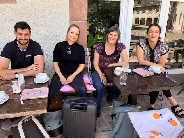 Vier Personen sitzen auf Stühlen an zwei kleinen Holztischen. Im Hintergrund sieht man eine steinerne Mauer sowie eine Glastür. Der Blick der Personen ist frontal in die Kamera gerichtet, sie lächeln. Auf dem Tisch stehen Tassen und Gläser und es liegen Notizblöcke darauf. Im Raum zwischen beiden Tischen steht ein Koffer und mehrere Rucksäcke.
