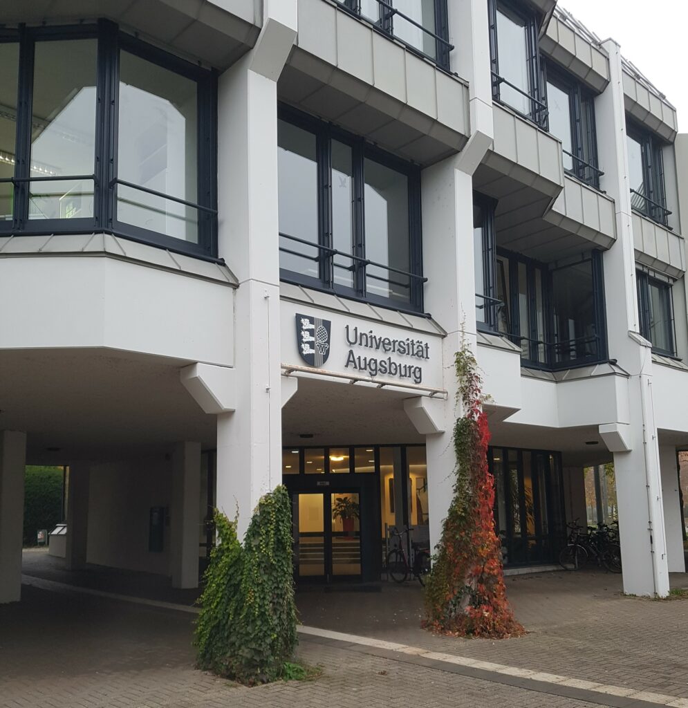 Das Bild zeigt ein zweistöckiges Gebäude mit einer grauen Fassade, in welcher zahlreiche Fenster mit schwarzem Rahmen eingelassen sind. In der Mitte über einem Durchgang steht der Schriftzug Universität Augsburg rechts neben dem Wappen der Universität. Zwei Sockel sind mit grünem und herbstlich rotem Efeu bewachsen und reichen fast bis hinaus zu dem Schriftzug.