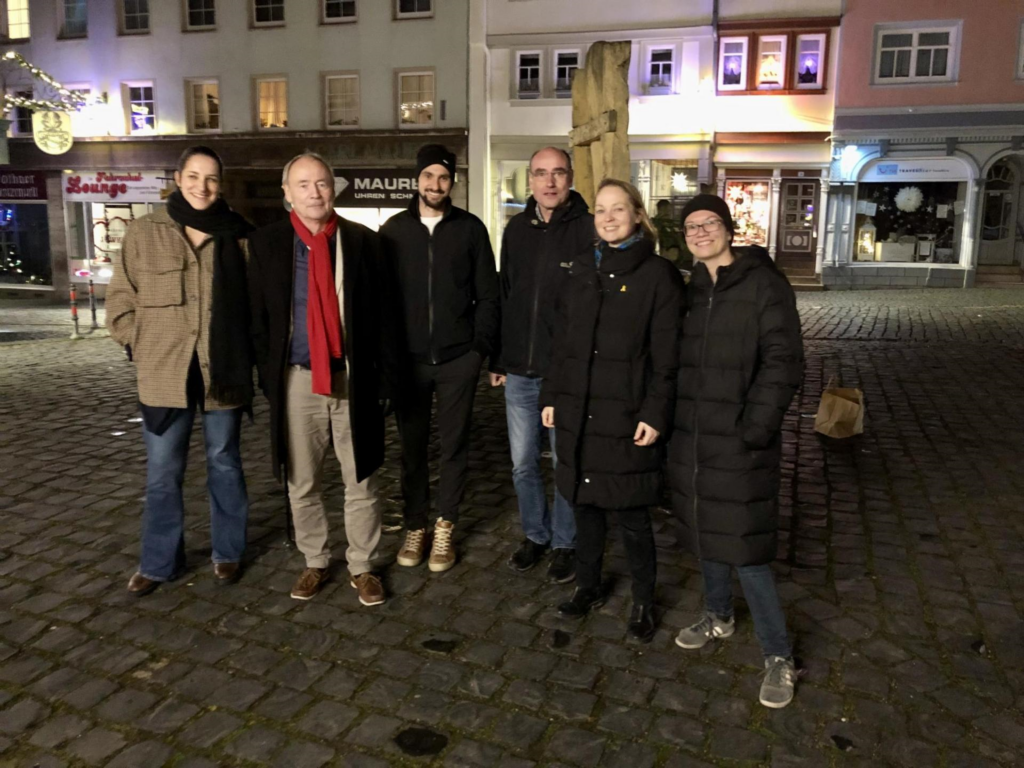 Auf dem Bild sind insgesamt sechs Personen zu sehen, die leicht seitlich gestellt in die Kamera blicken. Die Personen stehen auf einem Platz in der Innenstadt in Gotha auf Pflastersteinen. Dahinter sind beleuchtete Schaufenster zu sehen.